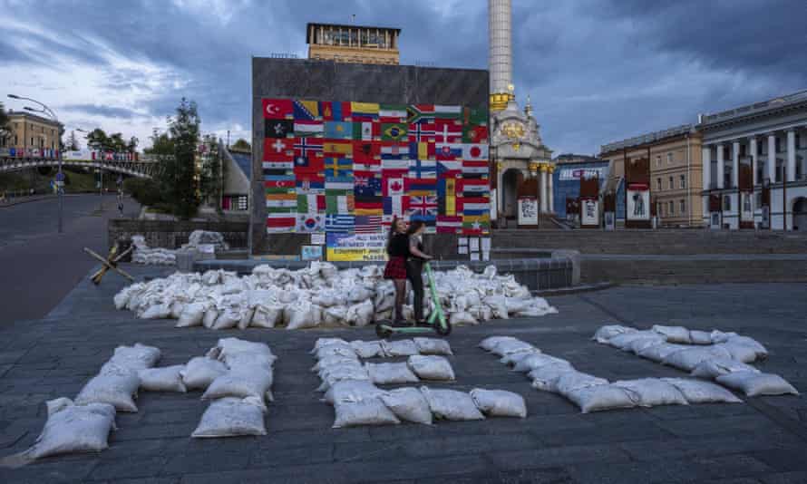 Women riding a motorcycle through Maidan Square in Kyiv, passing sandbags with the word 