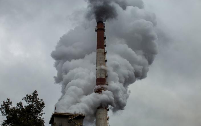 Viewed from under the overcast sky, smoke billows from the chimney of a red and white factory.
