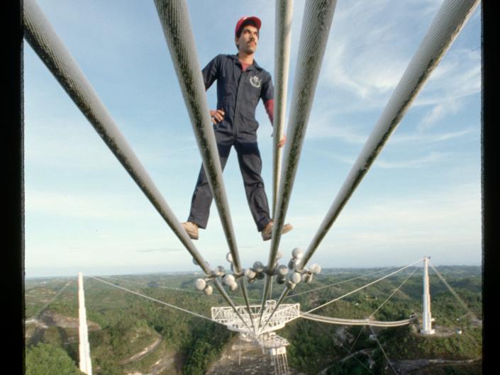 A man on cables above the ground above a radio telescope